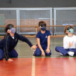 Três crianças estão sentadas no chão. São dois meninos e uma menina. Eles estão vendados a atividade é goalball.