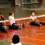 Duas alunas e a professora de educação física estão sentadas no chão em frente a uma rede de volêi. O esporte paralimpico é o vôlei sentado. As alunas estão com camisetas de uniformes brancas e a professora com a camiseta de uniforme azul marinho.