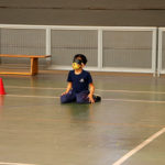 Fotografia de um menino sentado no chão da quadra de esportes com uma venda no olho. A atividade é a goalball. O menino está com o uniforem azul marinho do Colégio Miguel de Cervantes.