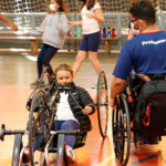 Uma aluna está sentada em uma handbike, bicicleta adaptada. Ao seu lado o professor André que é cadeirante e está em sua cadeira de rodas está orientando a aluna. A aluna está com os cabelos loiros amarrados e usa uma camiseta branca do uniforme do Colégio e uma jaqueta preta. O professor André está sentado de costas com a camiseta azul marinho do uniforme do Colégio. O professor André é negro, usa óculos e tem o cabelo curto enrolado. A aluna e o professor usam máscara de proteção.