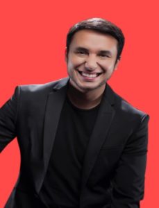 Retrato de Alex Duarte: homem branco de cabelo curto de cor castanho escuro. Rosto afilado, mas levemente redondo, nariz largo e boca grande. Ele, vestindo um blazer preto por cima de uma camiseta preta, está sorrindo largamente em frente a um fundo integralmente vermelho. Seu corpo está levemente inclinado para a esquerda e só é visto de sua cintura para cima. Seus braços estão dispostos de maneira que não mostra suas mãos.