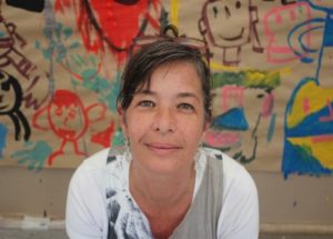 Retrato de Carla Mauch: mulher branca de cabelo escuro que na fotografia aparece preso com um par de óculos. Rosto arredondado, nariz fino e boca grande. Ela posa com uma blusa de cores cinza e branco, na qual as mangas são brancas, mostrando um sorriso tímido sem mostrar os dentes. Está em frente a um quadro com diversas pinturas coloridas feitas, aparentemente, por crianças. A fotografia é de seu pescoço para cima.