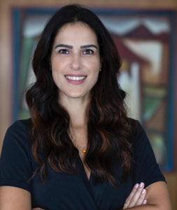 Retrato de Carolina Videira: mulher branca com cabelos longos ondulados de cor predominantemente castanho escuro, mas com mechas castanhas claras nas ondulações. Rosto fino e afilado, nariz fino e lábios grossos. Ela, vestindo uma blusa azul escura levemente decotada, posa com os braços cruzados em frente a um fundo desfocado, no qual se encontra uma pintura. É visto de seu peitoral para cima.