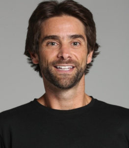 Retrato de Henry Zylberstajn: homem branco de cabelo cor castanho escuro de comprimento médio e barba e bigode ralos da mesma cor do cabelo. Rosto afilado, nariz e lábios finos. Em frente a um fundo integralmente branco, ele sorri largamente. Usa uma camiseta preta e é visto de seus ombros para cima.