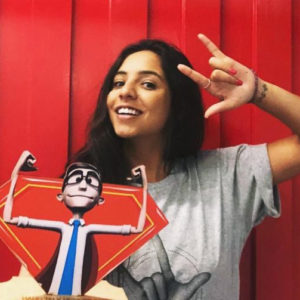 Retrato de Rafaella Franciscatto: mulher negra de cabelo liso longo de cor preta. Rosto afilado, nariz largo e lábios finos. Ela usa uma camisa cinza com a estampa de uma mão aberta com os dois dedos do meio abaixados e segura, com a mão direita, uma imagem de um homem branco, de óculos, usando uma gravata azul e uma capa de super-herói que faz o movimento que representa força, com os dois braços erguidos. Com a mão esquerda erguida, ela reproduz a estampa de sua camiseta, coincidentemente ou não. Ela tem as unhas pintadas de vermelho, um anel no dedo indicador da mão esquerda, uma pequena tatuagem e uma pulseira no punho esquerdo. Ela mostra um largo sorriso em frente a uma parede integralmente vermelha, tendo a cabeça levemente virada para a direita. A foto é do peitoral para cima.