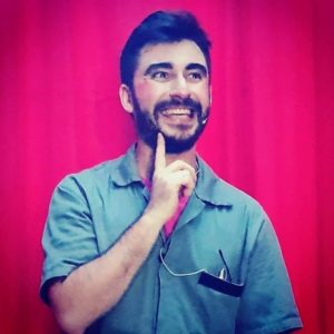 Retrato de Thiago Franco: homem branco de cabelo curto de cor castanho escuro e barba e bigode levemente cheios da mesma cor do cabelo. Rosto afilado, nariz e lábios finos. Usando uma camisa verde musgo, por cima de uma camisa vermelha, ele sorri amplamente em frente a algo que aparenta ser uma cortina de palco de cor vermelha. Ele carrega, no bolso frontal da camisa, um dispositivo móvel ligado a um microfone, que está para fora do bolso. A fotografia foi tirada de maneira levemente lateral, fazendo com que o lado direito esteja mais aparente que o esquerdo. Na foto, que o coloca do peitoral para cima, ele está com o braço esquerdo disposto naturalmente ao lado do tronco e o direito está dobrado, tendo o dedo indicado tocando o queixo.