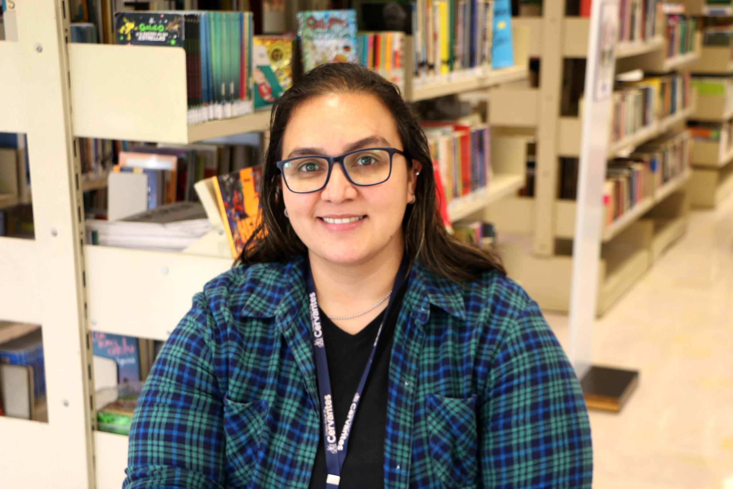 Retrato: A educadora está sentada em frente a uma estantes de livros. Ela temo o rosto redondo, usa óculos de aros  azuis, tem o cabelo castanho na altura dos ombros e está sorrindo. Ela está usando uma camiseta preta e por cima uma camisa xadrez azul, verde e preta. 